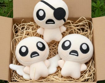 May include: Three plush figures with large black eyes and small arms are nestled in a cardboard box filled with straw. One figure has a black eye patch. The figures are a light beige color.
