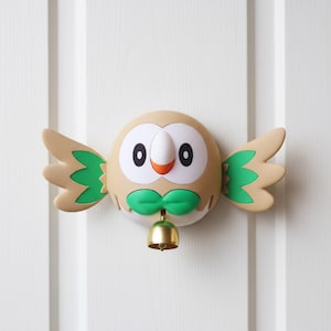 Könnte beinhalten: Eine dekorative Türklingel in Eulenform mit einem Körper in Beige, einem weißen Gesicht und grünen Akzenten. Die Eule hat große schwarze Augen, einen orangefarbenen Schnabel und eine grüne Fliege. Die Flügel sind beige mit grünen Details. Eine kleine goldene Glocke hängt darunter.
