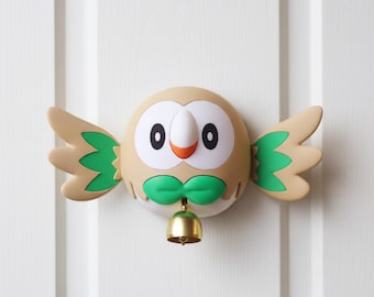 May include: A decorative owl-shaped doorbell with a tan body, white face, and green accents. The owl has large black eyes, an orange beak, and a green bow tie. The wings are tan with green details. A small gold bell hangs below.