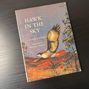 May include: A paperback book titled Hawk in the Sky by Franklin Russell, illustrated by Fredric Sweney. The cover features a painting of a hawk in flight over a landscape with orange and brown tones, with the text TW 3932 in the top corner.
