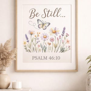 Può includere: Una stampa incorniciata con un'illustrazione ad acquerello di fiori selvatici e una farfalla. Il testo "Be Still..." e "Psalm 46:10" sono visualizzati in una scrittura elegante. L'opera d'arte è incorniciata in una cornice di legno chiaro.