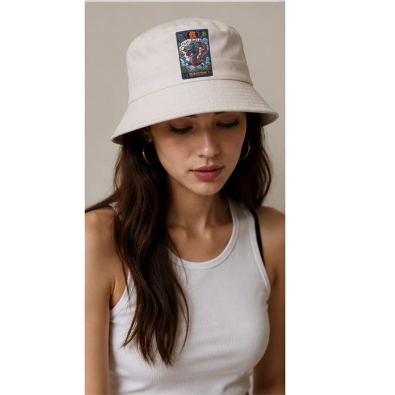 May include: Off-white bucket hat with a rectangular patch featuring a colorful illustration of a dragon. The hat has a wide brim and is made of a soft fabric. The word "DRAGON" is printed on the patch.