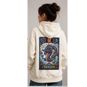 May include: Cream-colored hoodie with a colorful dragon graphic on the back. The dragon is depicted in shades of blue, green, and red, with the word "DRAGON" below. The hoodie has a drawstring hood and a kangaroo pocket.
