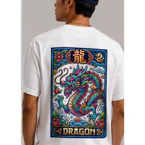 May include: White t-shirt featuring a colorful dragon design on the back. The dragon is depicted in shades of blue, green, and red, with the word "DRAGON" below. The design is framed by a blue border with floral and wave motifs.