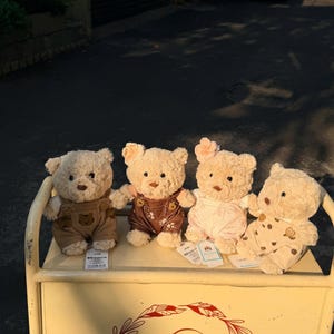 May include: Four plush teddy bears with varying outfits, including overalls and patterned rompers, are displayed on a cream-colored cart. The bears have light brown fur and are positioned in a row. The cart has a decorative logo.