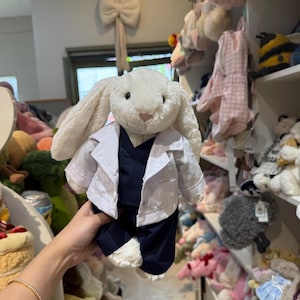 May include: A plush white bunny wearing a white lab coat and navy blue pants. The bunny has long, floppy ears and a pink nose. The bunny is being held in front of a store display filled with other stuffed animals.