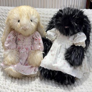 Puede incluir: Dos muñecas de peluche con forma de conejo. Una es de color crema con un vestido floral rosa. La otra es negra con un vestido floral blanco. Ambas muñecas tienen pelaje esponjoso y están sentadas sobre una superficie blanca texturizada.