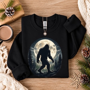 May include: A black crewneck sweatshirt featuring a circular graphic of a dark Bigfoot silhouette walking through a forest at night under a large, bright full moon. The garment has a Gildan brand label at the inner neckline.