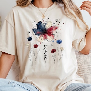 May include: Beige t-shirt featuring a watercolor floral design in red, white, and blue, with the word "america" in script. The design includes butterflies and stars, evoking a patriotic theme. The shirt is a relaxed fit.