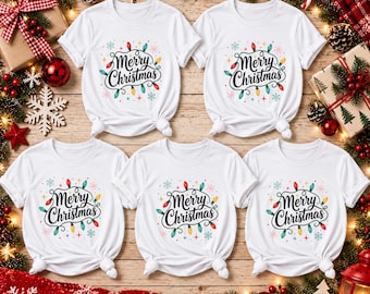 Puede incluir: Cinco camisetas blancas de manga corta dispuestas en grupo, cada una con un estampado gráfico del texto Merry Christmas en letra negra, rodeado de bombillas navideñas de colores e ilustraciones de copos de nieve azul claro.