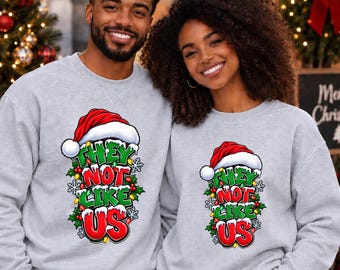 Puede incluir: Dos sudaderas de cuello redondo en color gris claro con un estampado gráfico de un gorro de Papá Noel rojo sobre texto verde y rojo que dice THEY NOT LIKE US, rodeado de hojas de acebo, bayas, luces navideñas y copos de nieve.