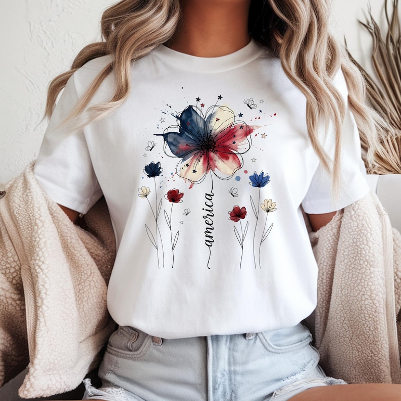 May include: White t-shirt featuring a watercolor floral design in red, white, and blue, with the word "america" in a cursive font. The design includes butterflies and stars, evoking a patriotic theme.