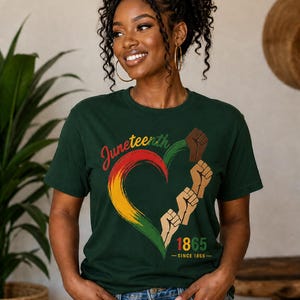May include: Dark green t-shirt featuring a heart-shaped design in red, yellow, and green, with the word "Juneteenth" in script. The design includes raised fists and the year "1865". The shirt is worn by a person with curly hair.