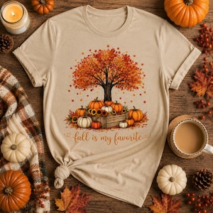 Puede incluir: Una camiseta beige de manga corta con un gr&aacute;fico de un &aacute;rbol grande con hojas de oto&ntilde;o naranjas y rojas, calabazas y girasoles en una caja de madera. El texto "fall is my favorite" est&aacute; impreso en una fuente cursiva debajo.