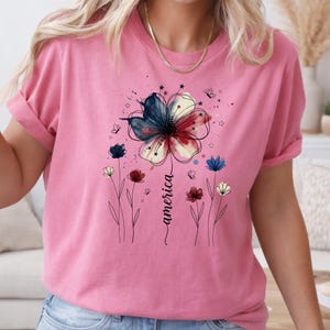 May include: Pink t-shirt with a floral design in red, white, and blue, with the word "america" in cursive. The design includes small flowers and butterflies. The shirt has rolled-up sleeves.