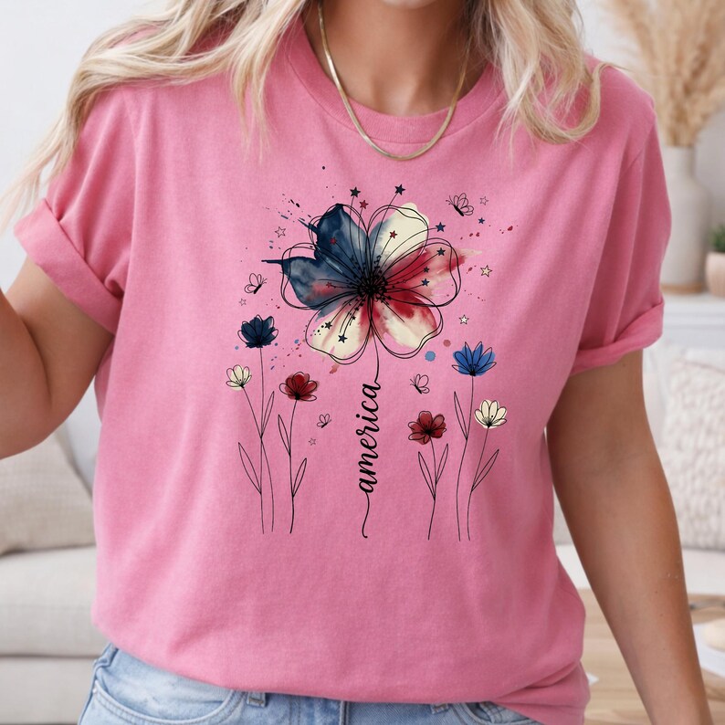 May include: Pink t-shirt with a floral design in red, white, and blue, with the word "america" in cursive. The design includes small flowers and butterflies. The shirt has rolled-up sleeves.