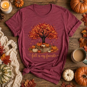Puede incluir: Una camiseta de manga corta color granate con un gr&aacute;fico de un &aacute;rbol con hojas de oto&ntilde;o naranjas y rojas sobre una caja de madera llena de calabazas. El texto "fall is my favorite" est&aacute; impreso en una fuente cursiva dorada debajo.