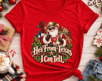 Puede incluir: Una camiseta roja de manga corta con un gráfico de Santa Claus con sombrero de vaquero y gafas de sol, rodeado de cactus, un molino de viento y la bandera de Texas. El texto dice "He's From Texas I Can Tell" en una fuente decorativa.
