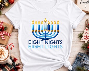 Puede incluir: Una camiseta blanca de manga corta con un gráfico de una menorá azul de nueve brazos con llamas amarillas encendidas. Debajo de la menorá, el texto EIGHT NIGHTS EIGHT LIGHTS aparece en letras mayúsculas azules y azul claro.
