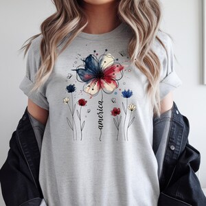May include: A light grey t-shirt with a floral design in red, white, and blue, and the word "america" in a script font. The design includes a large flower and smaller flowers.