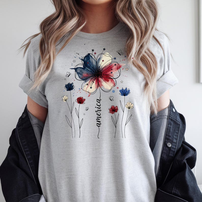 May include: A light grey t-shirt with a floral design in red, white, and blue, and the word "america" in a script font. The design includes a large flower and smaller flowers.