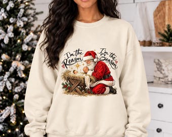 Puede incluir: Una sudadera de cuello redondo color crema con un estampado gráfico de Santa Claus arrodillado junto a un pesebre con un bebé. El texto "I'm the Reason" y "I'm the Season" está impreso en negro sobre la ilustración, decorado con pequeños corazones rojos.