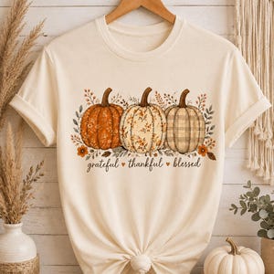 May include: A cream-colored short-sleeve t-shirt featuring a graphic of three pumpkins with floral and plaid patterns. The text "grateful thankful blessed" is printed in black cursive script below the pumpkins, accented by small orange hearts.