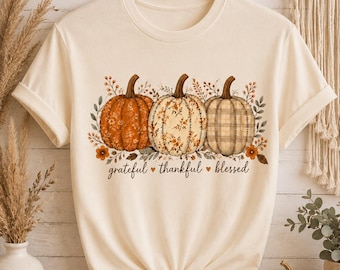 Puede incluir: Una camiseta de manga corta color crema con un gráfico de tres calabazas con patrones florales y de cuadros. El texto "grateful thankful blessed" está impreso en cursiva negra debajo de las calabazas, acentuado por pequeños corazones naranjas.