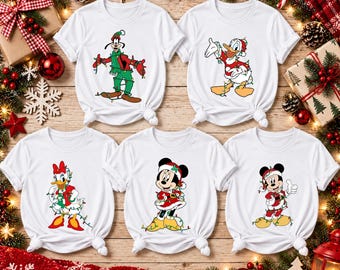 Puede incluir: Cinco camisetas blancas de manga corta con los personajes de Disney Goofy, el Pato Donald, Daisy, Minnie Mouse y Mickey Mouse. Cada personaje viste ropa festiva y está envuelto en guirnaldas de luces navideñas de colores.