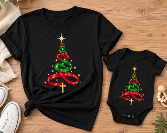 Puede incluir: Una camiseta negra de manga corta y un body de bebé negro a juego de manga corta. Ambas prendas presentan un gráfico de un árbol de Navidad formado por cintas rojas y verdes, decorado con estrellas amarillas y una cruz dorada en la base.