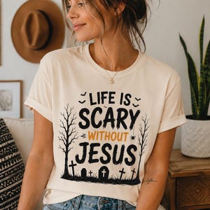 May include: A cream-colored short-sleeve t-shirt featuring a black and orange graphic print. The text reads "LIFE IS SCARY WITHOUT JESUS" in a distressed font, surrounded by illustrations of bare trees, tombstones, bats, and a small spider.