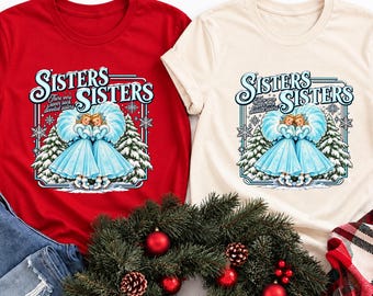 Puede incluir: Una camiseta roja y una camiseta color crema, cada una con un gráfico de dos figuras con vestidos azules con el texto "Sisters Sisters" y "There were never such devoted sisters" rodeado de copos de nieve y pinos nevados.