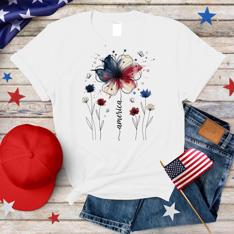 May include: White t-shirt with a watercolour floral design in red, white, and blue, with the word "america" in script. The design includes stars and butterflies. A red baseball cap and American flag are also visible.