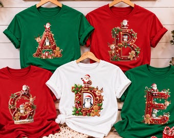Puede incluir: Cinco camisetas de manga corta en rojo, verde y blanco, cada una con una letra grande y ornamentada con temática navideña. Las letras A, B, G, D y E están decoradas con Papá Noel, renos, pingüinos, hombres de jengibre y acebo.