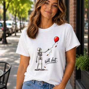 May include: A white short-sleeve t-shirt featuring a graphic of a skeleton reaching for a floating red balloon. The text "Well, shit..." is printed in black script below the skeleton, accompanied by small brown falling leaves.