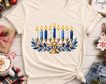 Puede incluir: Una camiseta de color crema con un gráfico de una menorá dorada con nueve velas encendidas en tonos alternos de azul y blanco. La menorá está decorada con hojas azules y una Estrella de David en la base, centrada en la parte frontal.