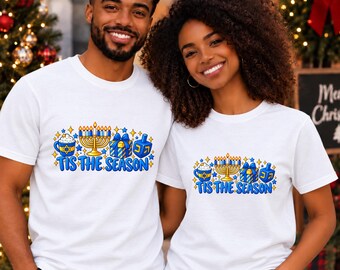 Puede incluir: Una camiseta blanca de manga corta con un estampado gráfico de una menorá azul y dorada, un regalo, una peonza dreidel y una taza con una estrella de David. El texto 'TIS THE SEASON' está impreso en azul debajo de las ilustraciones.