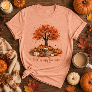 Puede incluir: Una camiseta color melocot&oacute;n con el gr&aacute;fico de un &aacute;rbol con hojas de oto&ntilde;o naranjas y rojas sobre una caja de madera llena de calabazas y girasoles. El texto "fall is my favorite" est&aacute; impreso en una fuente cursiva marr&oacute;n bajo la caja.