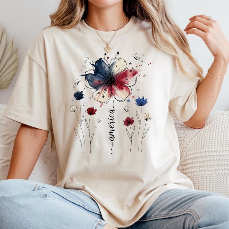 May include: Beige t-shirt featuring a watercolor floral design in red, white, and blue, with the word "america" in script. The design includes butterflies and stars, evoking a patriotic theme. The shirt is a relaxed fit.