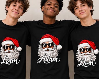 Puede incluir: Tres camisetas negras con un gráfico de Santa Claus con gafas de sol negras y un gorro rojo. Cada camiseta muestra un nombre único en letra cursiva blanca debajo del gráfico: Liam, Adam y Noah, impresos en la parte frontal de las prendas.