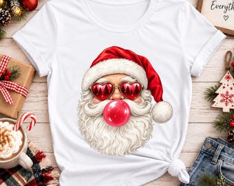 Puede incluir: Una camiseta blanca de manga corta con un gráfico de Santa Claus con un gorro rojo y gafas de sol en forma de corazón con estampado de bosque. Santa tiene una gran barba blanca y está inflando una burbuja de chicle roja.