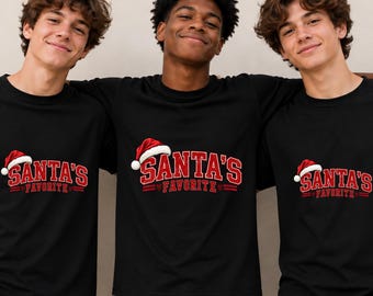Puede incluir: Una camiseta negra de cuello redondo con un estampado gráfico en rojo y blanco que dice SANTA'S FAVORITE en una fuente universitaria en negrita. El diseño incluye un gorro de Papá Noel rojo y blanco sobre la letra S y corazones rojos.