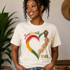 May include: Cream-colored t-shirt with a heart-shaped design in red, yellow, and green, featuring raised fists and the text "Juneteenth 1865". The shirt is a celebration of freedom and heritage.