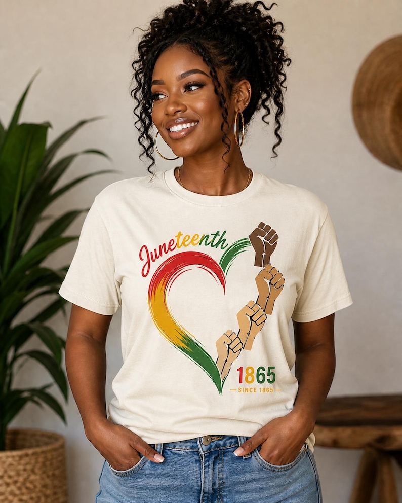 May include: Cream-colored t-shirt with a heart-shaped design in red, yellow, and green, featuring raised fists and the text "Juneteenth 1865". The shirt is a celebration of freedom and heritage.