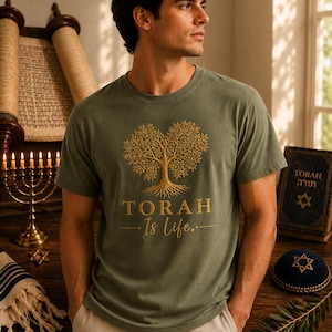 May include: An olive green short-sleeved t-shirt featuring a gold graphic of a tree with exposed roots. Below the tree, the text reads "TORAH" in a bold serif font, followed by the script text "Is life." centered between two horizontal lines.
