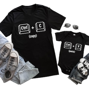 May include: Black t-shirts with white text. One shirt has "Ctrl + C (copy)" and the other has "Ctrl + V (paste)". Grey trainers and sunglasses are also in the image. The shirts are paired with blue jeans.
