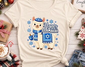 Puede incluir: Una camiseta color crema con el gráfico de una llama blanca que lleva un gorro y una bufanda de punto azul y blanco. La llama sostiene un dreidel azul. El texto dice Happy LLAMAKAH, rodeado de copos de nieve azules y una estrella de David.
