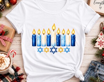 Puede incluir: Una camiseta blanca de manga corta con un gráfico de nueve velas encendidas en tonos azules y blancos, dispuestas como una menorá. Debajo de las velas hay cinco estrellas de David azules y amarillas centradas en el pecho.