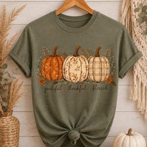 May include: An olive green t-shirt featuring a graphic of three pumpkins with different patterns, including floral and plaid designs. Below the pumpkins, the text reads "grateful thankful blessed" in a cursive font, with small orange heart icons.
