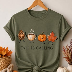 May include: An olive green short-sleeve t-shirt featuring a graphic of a smiling pinecone, a steaming coffee mug, a pumpkin, and a maple leaf. The text "FALL IS CALLING" is printed in black below the illustrations. The shirt has a crew neckline.